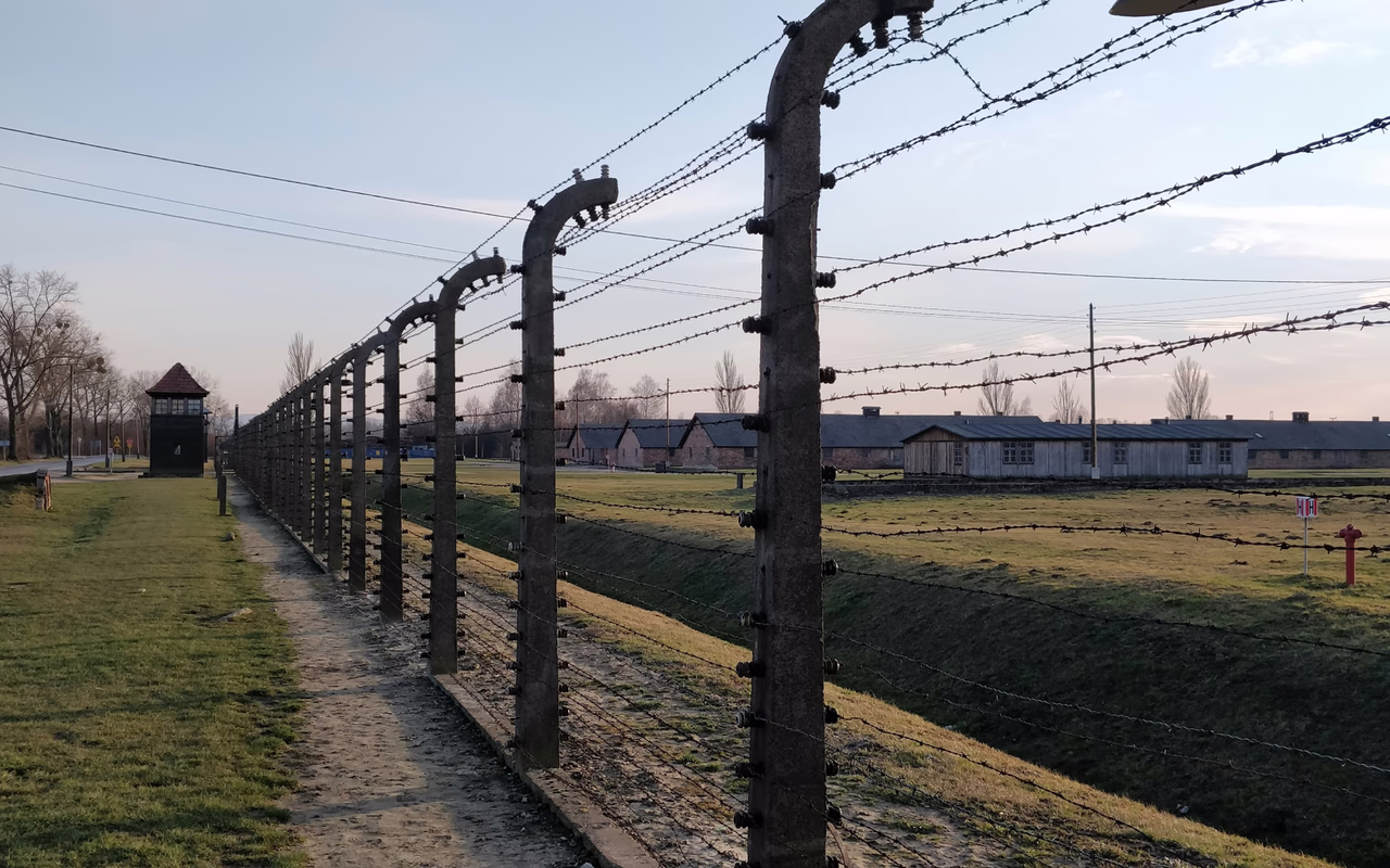 Bild eines Zauns im KZ Auschwitz (Oświęcim, Polen) mit einzelnen Häusern im Hintergrund